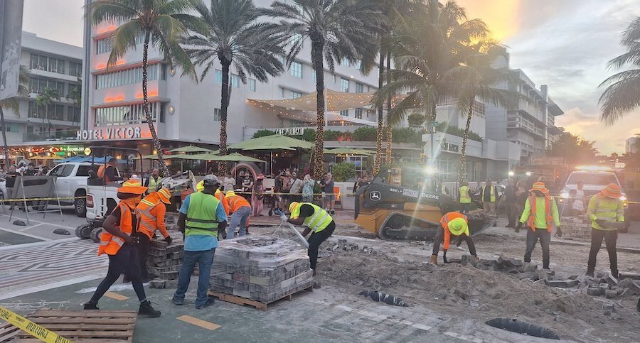Trabajadores con chalecos reflectivos y maquinaria pesada remueven los adoquines del cruce peatonal arcoíris en Ocean Drive, frente al Hotel Victor, al atardecer. Varias personas observan la escena desde las aceras. Crédito: Foto de Marina Romanello, publicada en X.